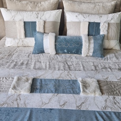 Almofadas e manta decorativas em tons de azul, branco e bege sobre cama com estofado bege.