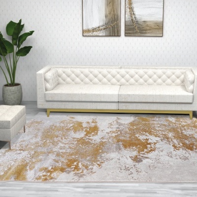 Sala de estar com sofá branco acolchoado, banco estofado, planta verde, tapete cinzento e dourado e pinturas abstratas na parede.