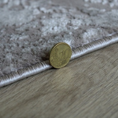 Moeda de 20 cêntimos junto à borda de tapete cinzento num chão de madeira.