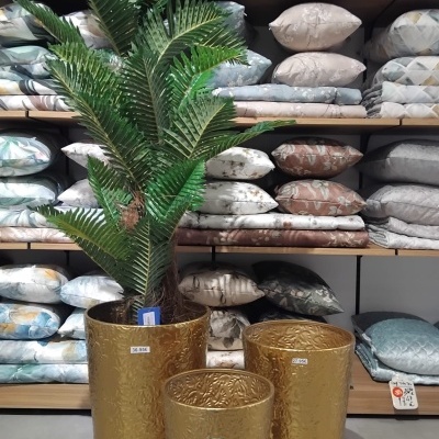 Três vasos dourados com planta artificial e prateleiras com almofadas e mantas