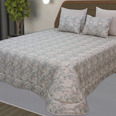 Colcha de cama com padrão floral azul e bege, quatro almofadas, cama com cabeceira castanha, planta e cortinas cinza.