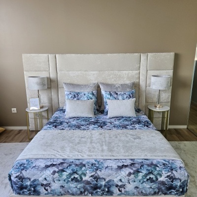 Cama com cabeceira cinza, edredão floral azul e cinzento com duas mesas de cabeceira douradas e lâmpadas cinzentas
