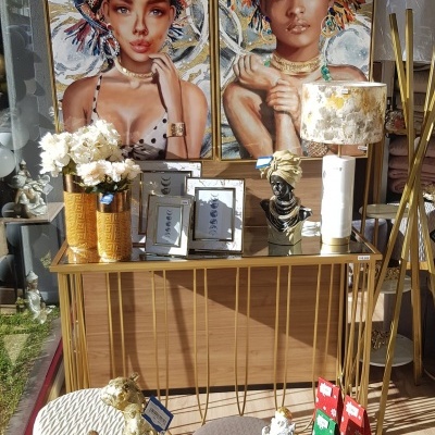 Área de exposição com quadros, móveis dourados, flores brancas, candeeiro e esculturas decorativas.