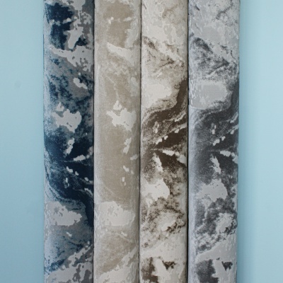 Tapetes enrolados com padrões abstratos em azul, bege, castanho e cinza encostados numa parede azul clara.