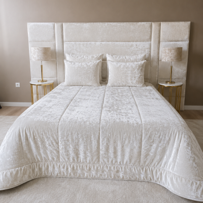 Cama de casal com edredão e almofadas bege claro em tecido veludo, com cabeceira estofada, duas mesas de cabeceira com candeeiros, parede castanha e chão de madeira clara.