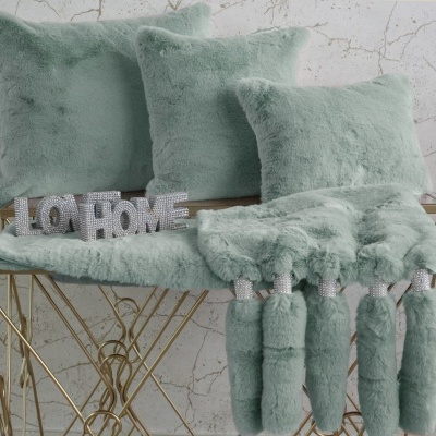 Três almofadas verdes de pelúcia e manta verde com franjas sobre mesa dourada com placa LO HOME