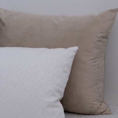 Duas almofadas, bege aveludada e branca com padrão em relevo, numa cama com roupa branca