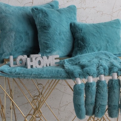 Almofadas e manta peluda azul-turquesa com tasséis em mesa dourada moderna.