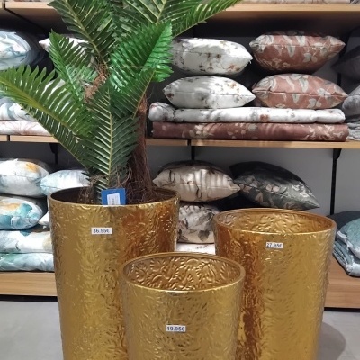 Três vasos dourados em metal com relevo, preços visíveis, um com planta artificial verde, almofadas florais em prateleiras ao fundo.