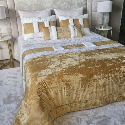 Cama com cabeceira estofada cinza, edredão e almofadas em veludo dourado e branco, mesa de cabeceira com abajur