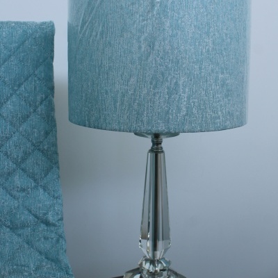 Abajur com base em vidro facetado e cúpula azul clara ao lado de cadeira azul estofada