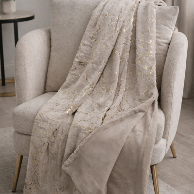 Cobertor bege com padrão dourado sobre poltrona bege em ambiente com plantas.