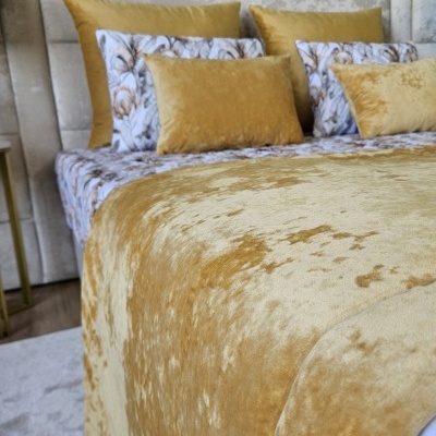 Cama com colcha e almofadas douradas e floridas com cabeceira cinza clara.