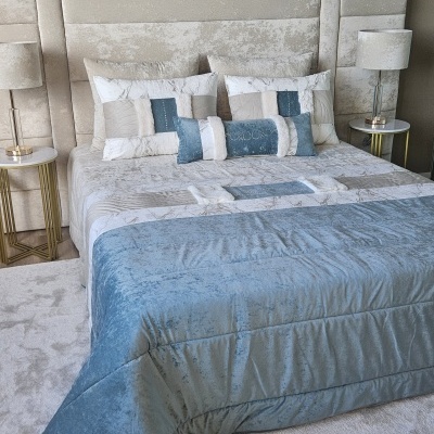 Cama estofada com colcha azul e branca e almofadas decorativas, com duas mesas de cabeceira e candeeiros