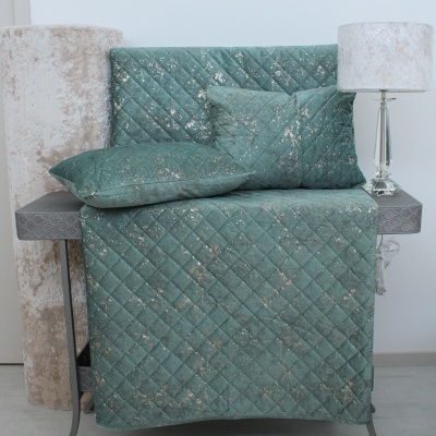 Conjunto de capa acolchoada e almofadas verde-azulado com padrão de diamante e manchas prateadas sobre móvel cinzento e candeeiro com abajur branco.