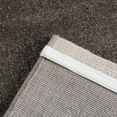 Canto de tapete castanho claro com borda bege sobre carpete castanho escuro.