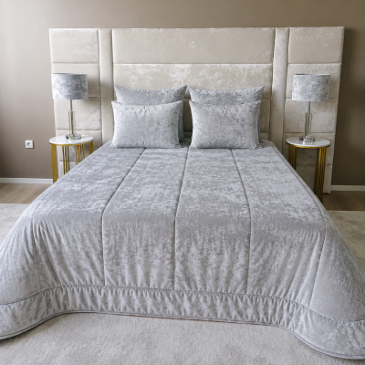 Quarto com cama de casal estofada bege, edredão cinza claro e duas lâmpadas com abajur cinza em mesas laterais brancas.