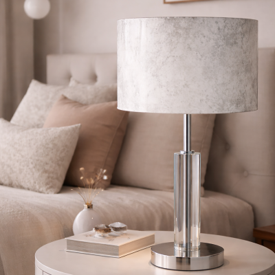 Candeeiro de mesa com base transparente e abat-jour cinza claro numa mesa branca junto a cama