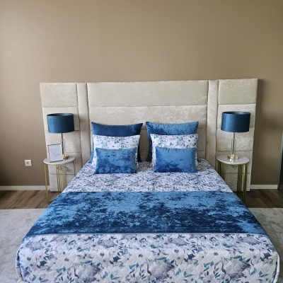 Cama com cabeceira estofada bege, roupa de cama floral e almofadas azuis, mesas laterais douradas, candeeiros azuis, parede castanha.
