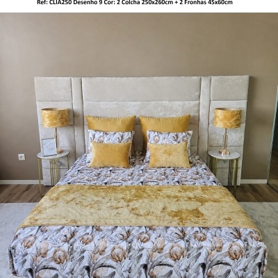 Cama estofada cinza claro com roupa de cama floral bege e castanho, almofadas amarelas, duas mesas de cabeceira com lâmpadas amarelas