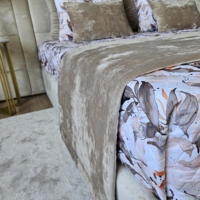Cama estofada bege com almofadas castanhas e padrão de folhas, edredão castanho claro e tapete bege.