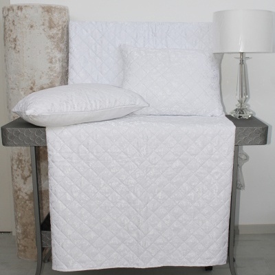 Conjunto de cama branco acolchoado com padrão em losango sobre mesa cinza ao lado de candeeiro de mesa