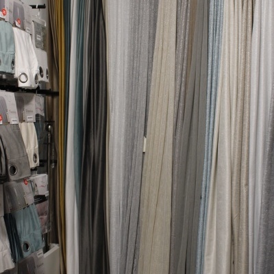 Várias cortinas em diferentes cores e texturas penduradas numa loja ao lado de prateleiras com amostras de tecidos.