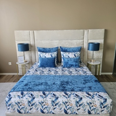 Cama de casal com cabeceira estofada bege e roupa de cama azul com padrão floral, almofadas azuis, duas mesinhas de cabeceira com abajures azuis.