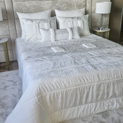 Cama com conjunto branco e cabeceira estofada cinza claro, almofadas brancas e mesas de apoio com candeeiros.