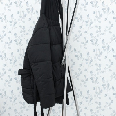 Casaco preto pendurado em cabide metálico prateado contra parede com padrão de folhas cinza claro