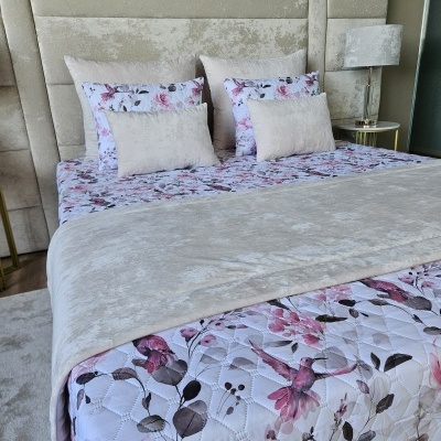 Cama com roupa de cama floral e almofadas beije com cabeceira estofada e candeeiro ao lado.