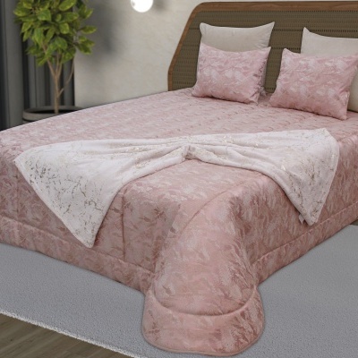 Cama com edredão rosa e almofadas, manta branca com padrão dourado, tapete cinza e planta interior