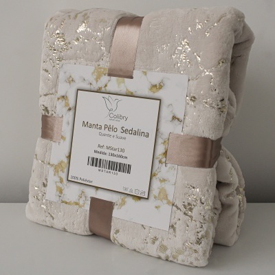 Manta de pelo bege com detalhes dourados, enrolada com fita castanha e etiqueta com texto Colibry Manta Pêlo Sedalina
