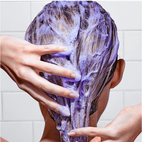 Cabelo com espuma roxa a ser aplicado por mãos sobre fundo de azulejos brancos