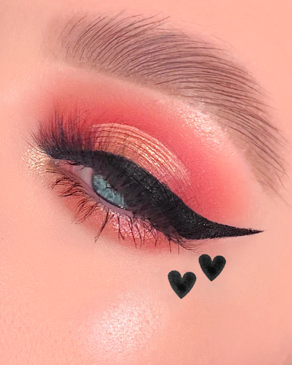 Olho maquilhado com sombra dourada e rosa, eyeliner preto e dois corações pretos desenhados na pele.