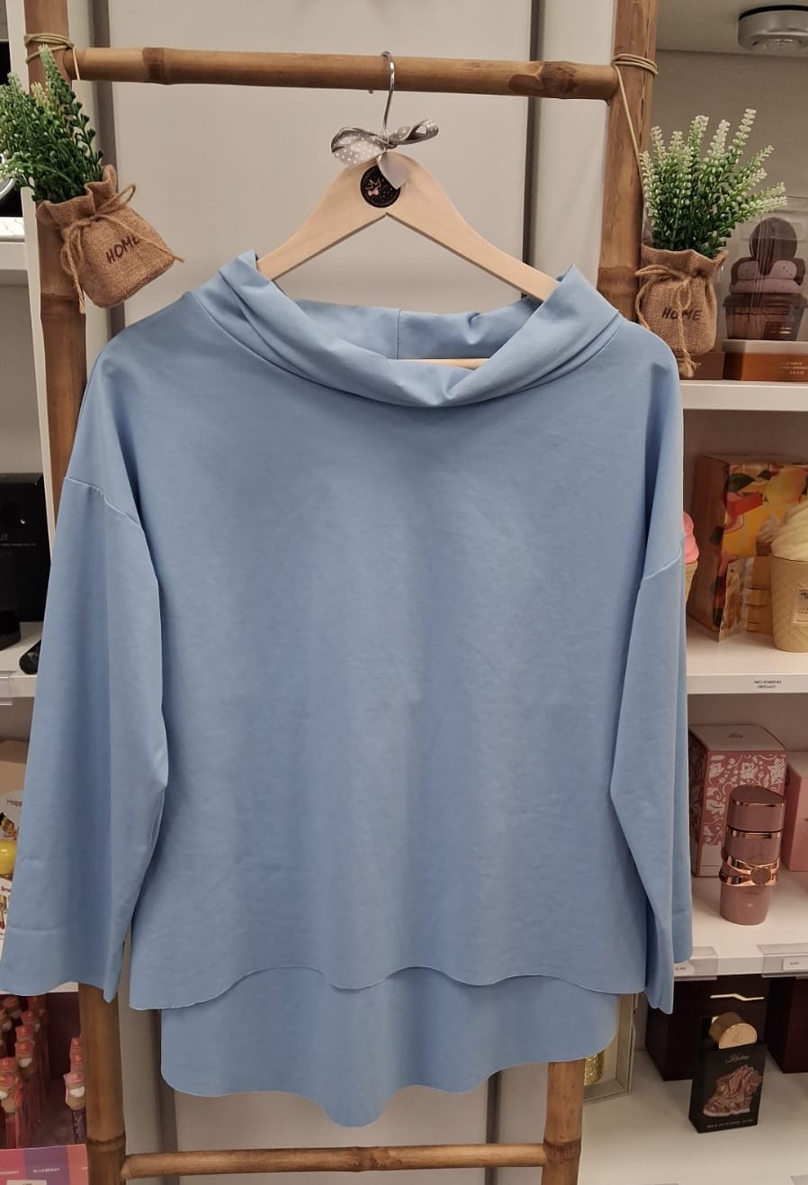 Camisola azul clara pendurada num cabide de madeira com fundo de estantes e plantas decorativas