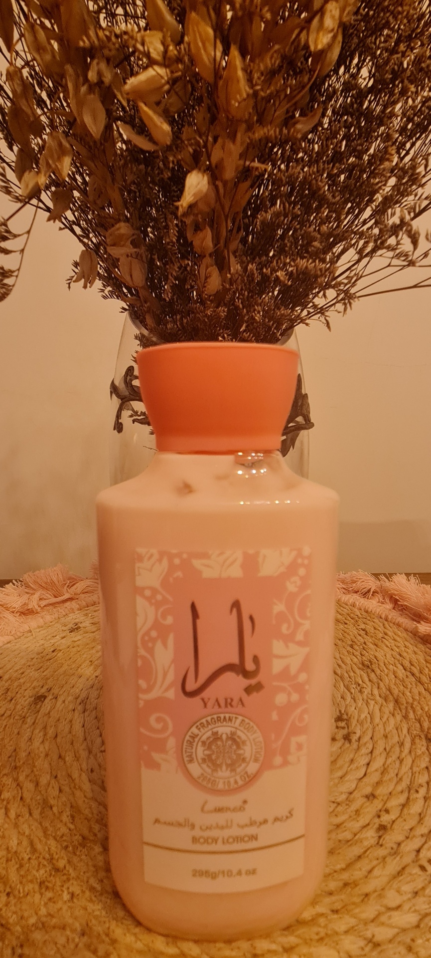 Frasco de hidratante corporal rosa com rótulo e tampa laranja junto a um vaso com flores secas.