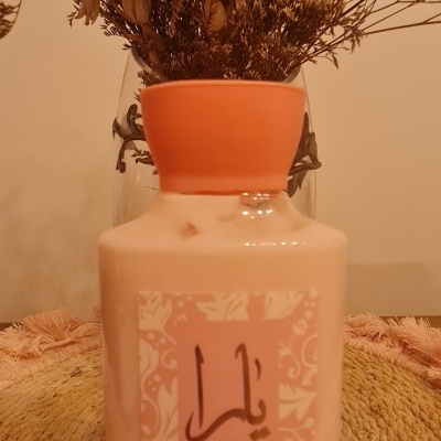 Frasco de hidratante corporal rosa com rótulo e tampa laranja junto a um vaso com flores secas.