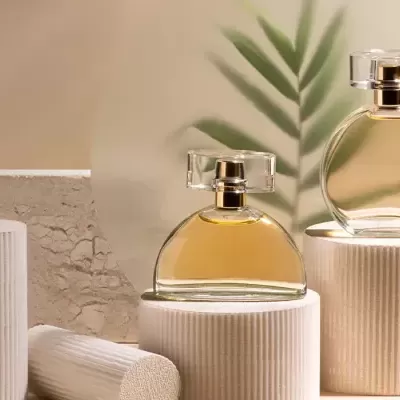 Dois frascos de perfume dourados em suportes brancos com embalagem e folhas decorativas ao fundo