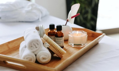 Tabuleiro de madeira com toalhas brancas, garrafas âmbar, escova, vela acesa e saco branco com laço vermelho