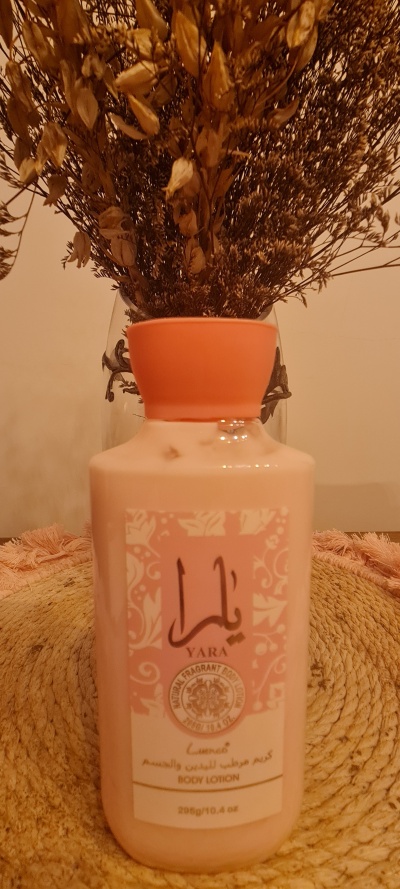 Frasco de hidratante corporal rosa com rótulo e tampa laranja junto a um vaso com flores secas.