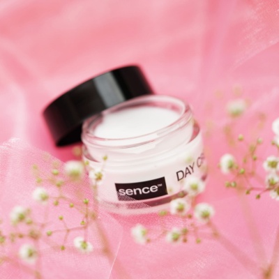 Frasco de creme facial com rótulo 'sence' e 'DAY C' sobre tecido rosa com flores brancas