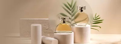Dois frascos de perfume dourados em suportes brancos com embalagem e folhas decorativas ao fundo