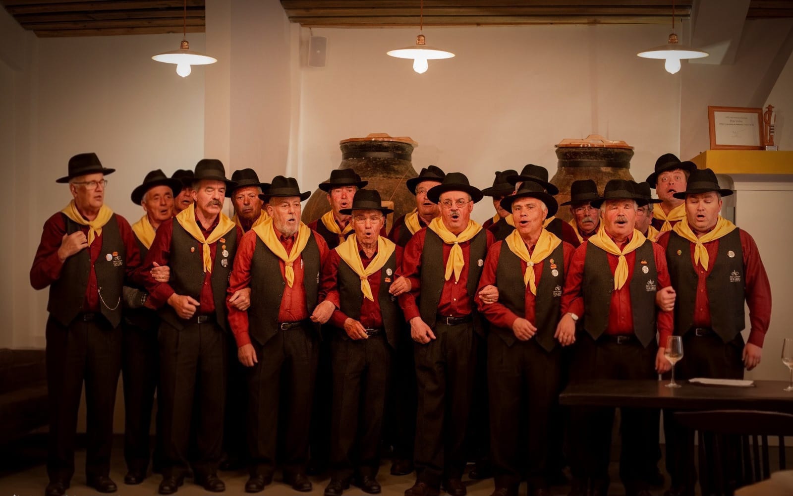 Grupo de homens com trajes tradicionais a cantar em interior iluminado.