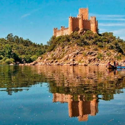 Castelo castanho-avermelhado numa colina refletido em água calma com vegetação e barco perto