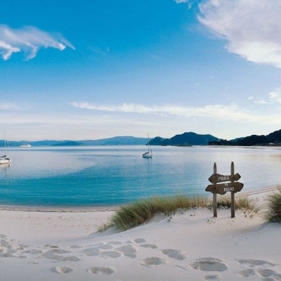 Praia com areia branca, vegetação rasteira, água azul calma e letreiro de madeira