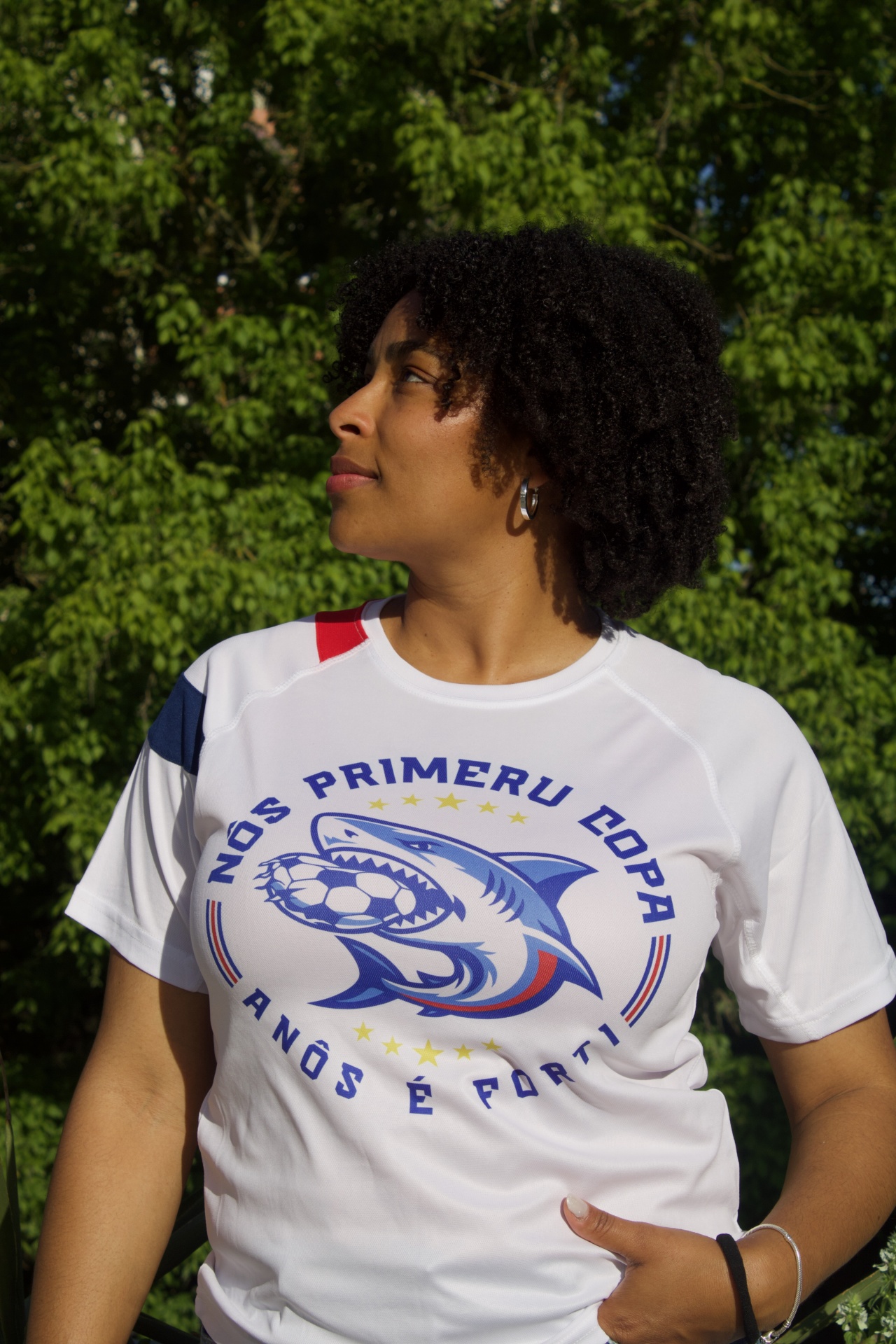 T-shirt branca com estampa de tubarão e texto em português vista em pessoa com cabelo encaracolado e fundo de árvores