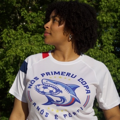 T-shirt branca com estampa de tubarão e texto em português vista em pessoa com cabelo encaracolado e fundo de árvores