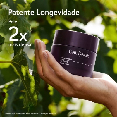 Frasco de creme roxo escuro CAUDALÍE Premier Cru segurado com fundo de folhas verdes