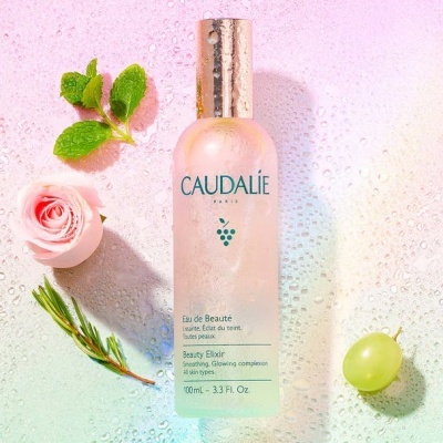 Frasco de Beauty Elixir da Caudalie com rosa, ramo verde e uva sobre superfície molhada